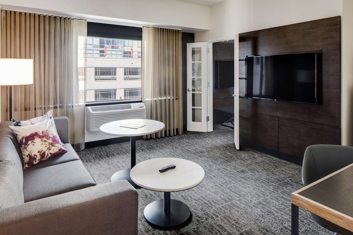 A hotel room with a sofa, a round table, a TV mounted on the wall, and a window with curtains.