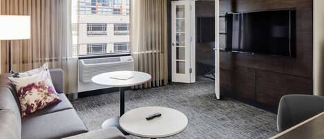 A hotel room with a sofa, a round table, a TV mounted on the wall, and a window with curtains.