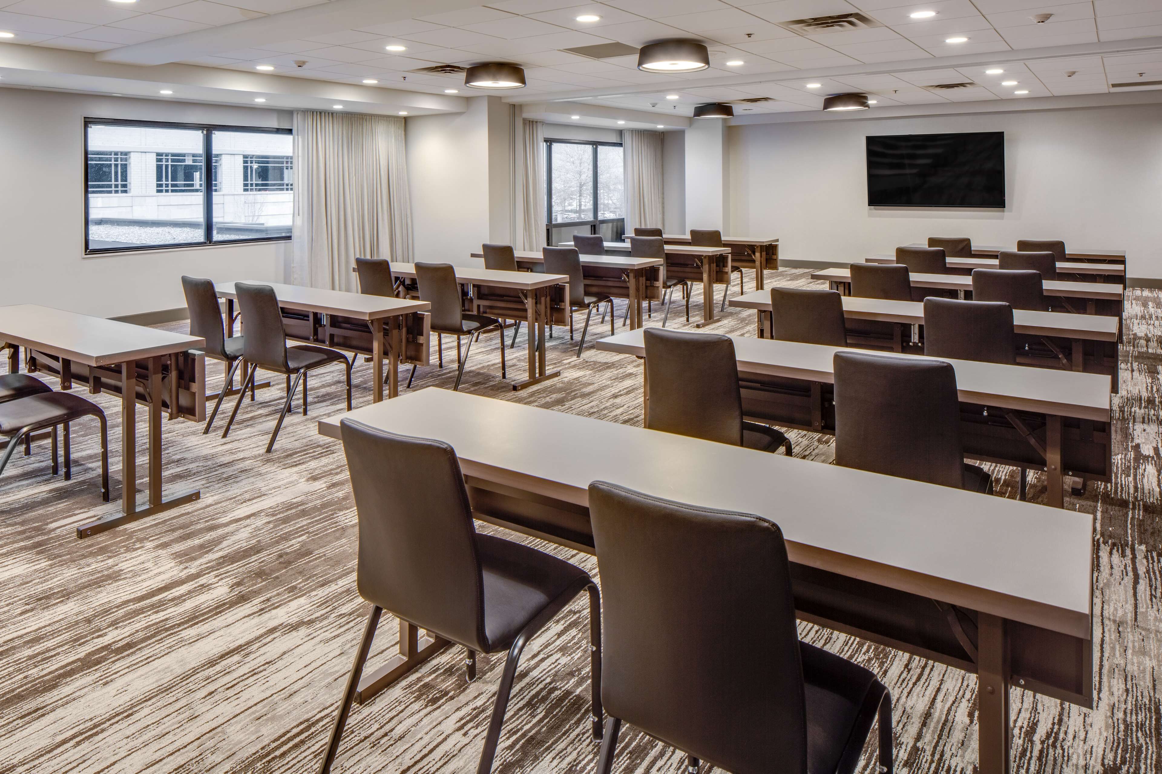 A conference room with multiple tables and chairs, large windows with curtains, and a mounted screen on the wall.