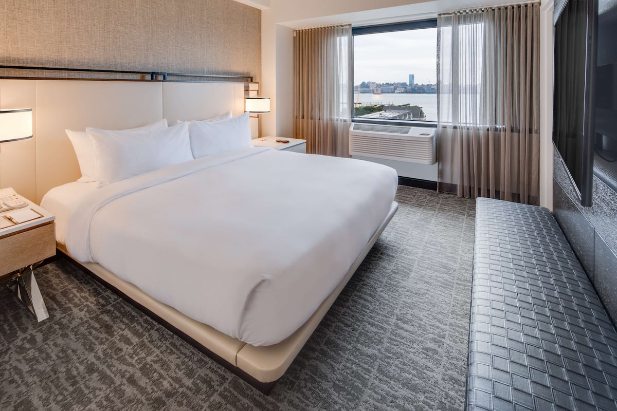 A hotel room with a large bed, a nightstand, a lamp, a window with curtains, and a view of a cityscape.