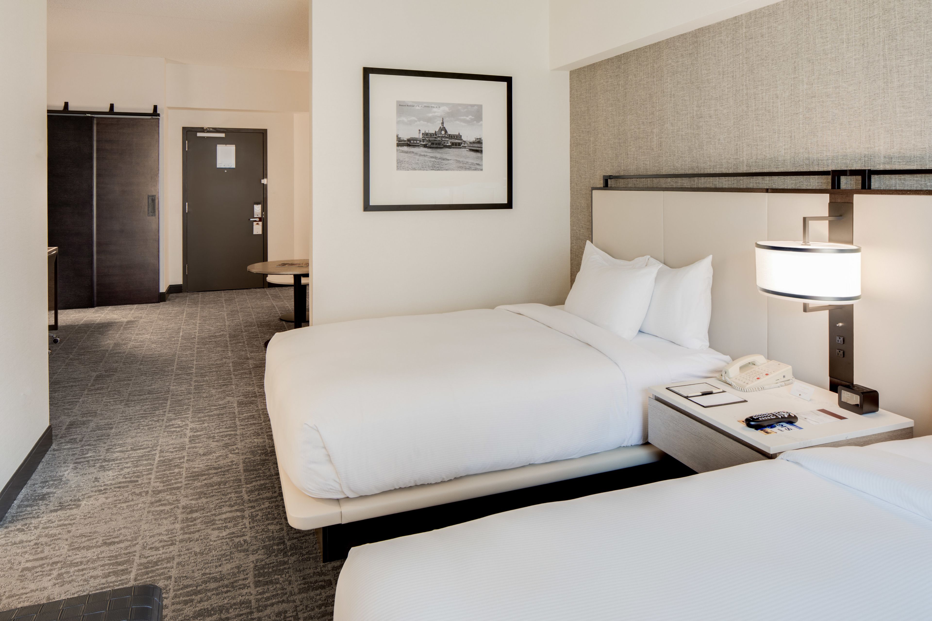 Habitación de hotel con dos camas, un cuadro enmarcado en la pared, una mesita de noche con lámpara y una puerta que da a otra habitación.