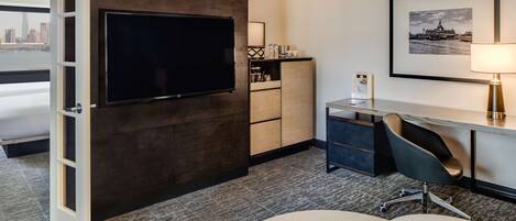 A hotel room with a large flat-screen TV mounted on a dark wall, a desk with a chair, a lamp, and a framed picture on the wall.