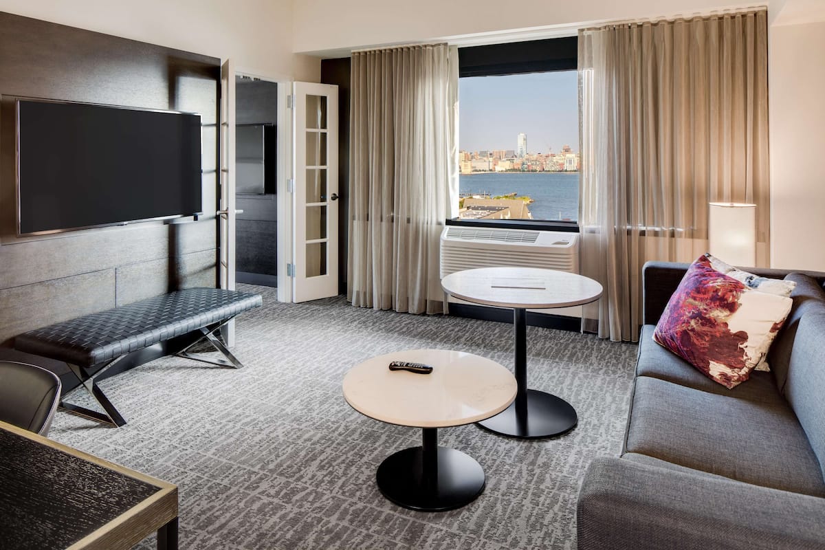 A hotel room with a flat-screen TV, a sofa, a bench, a small round table, and a view of a cityscape through large windows.