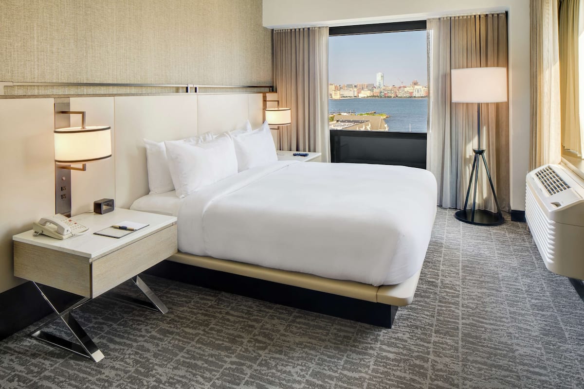 A hotel room with a large bed, a desk with a telephone, a lamp, and a view of the cityscape through the window.