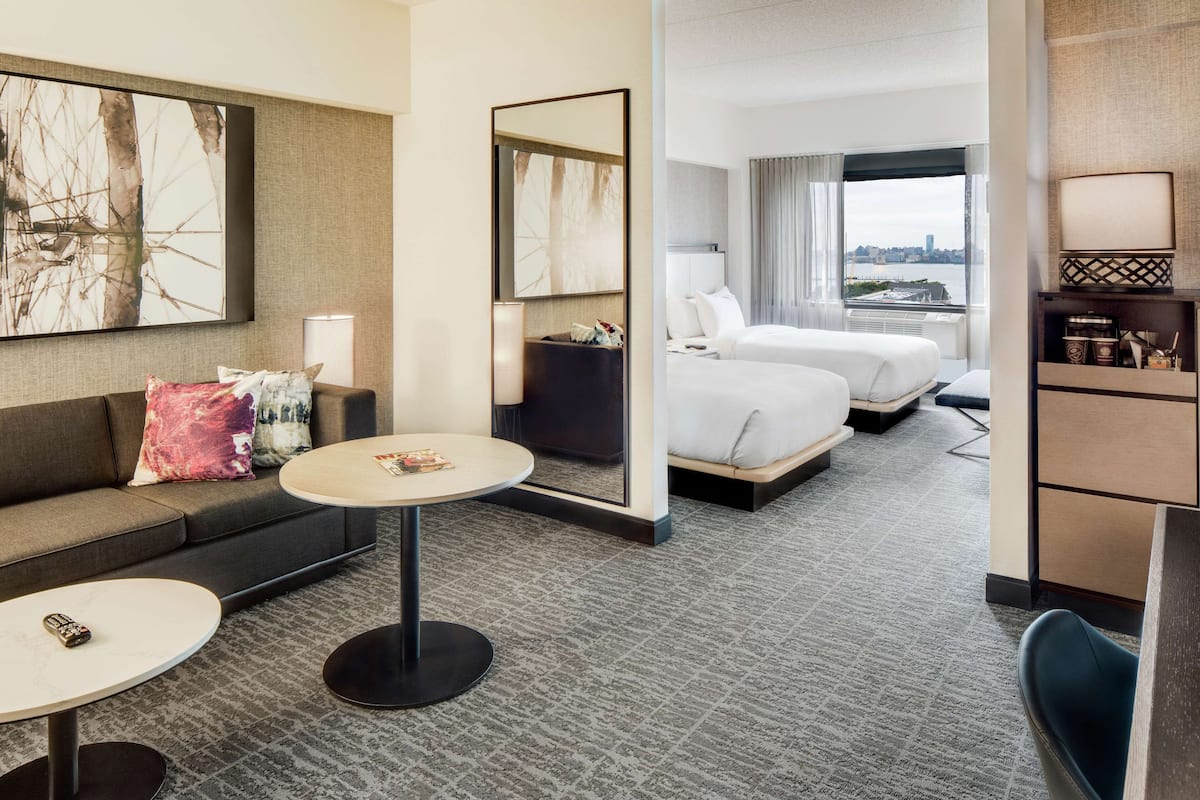A hotel room with a sofa, two round tables, a bed, a mirror, and a view of the city.
