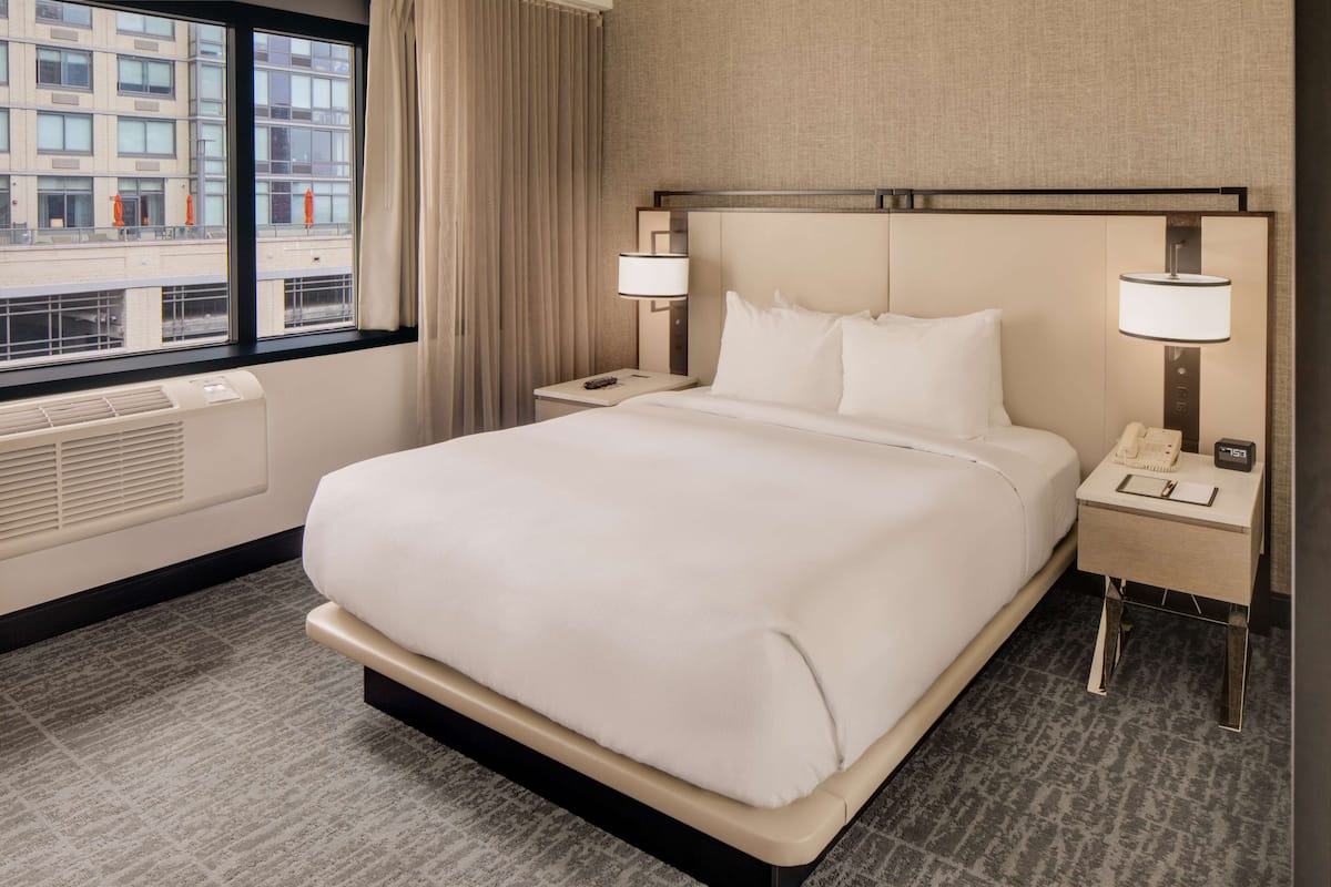 A hotel room with a large bed, two bedside tables with lamps, a window with curtains, and a view of buildings outside.