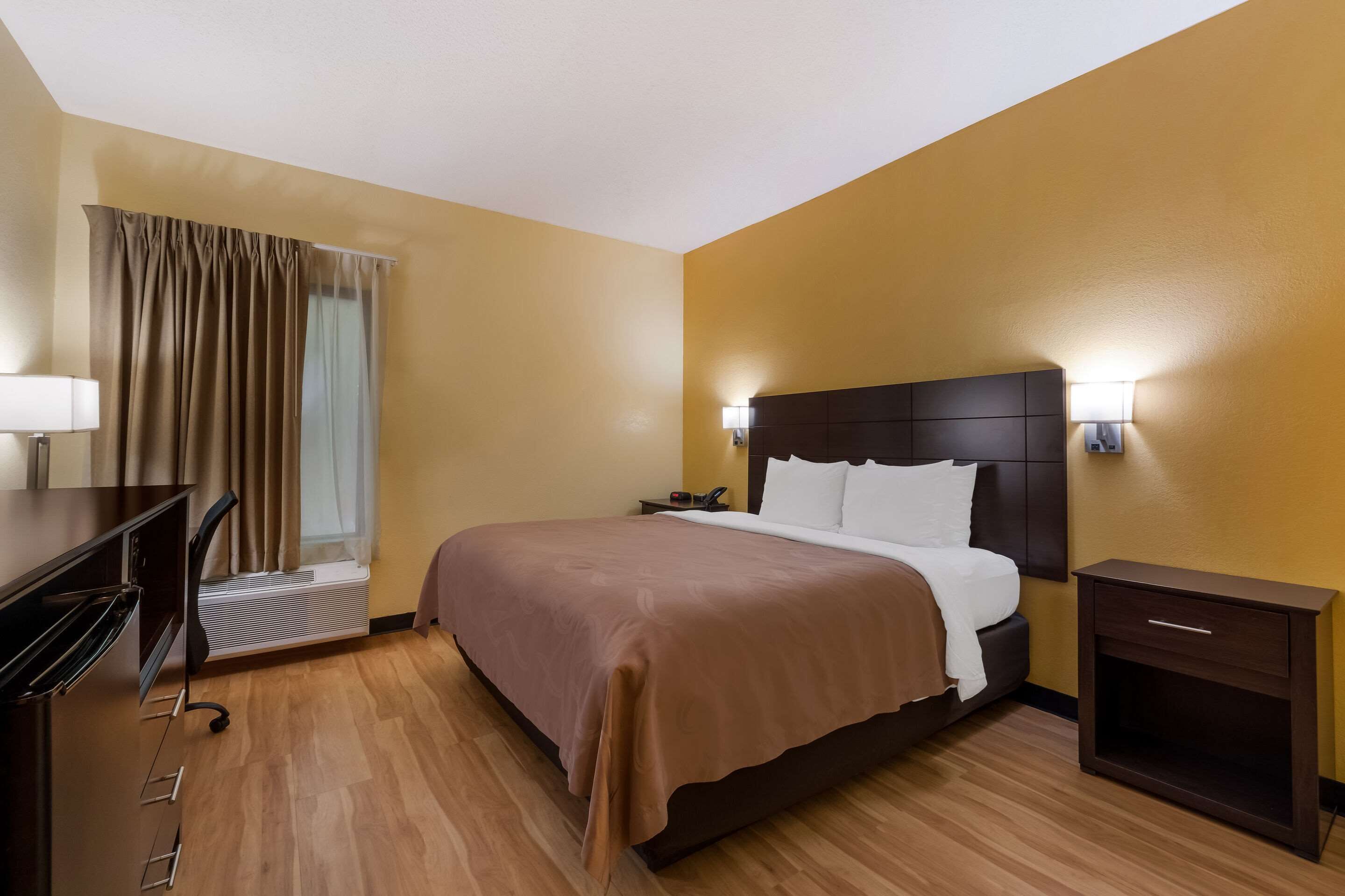 A hotel room with a bed, bedside table, lamp, and a desk with a chair.