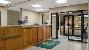 Un área de recepción con mostrador de madera, un letrero que dice “Quality Inn” y una puerta de cristal que da al exterior.