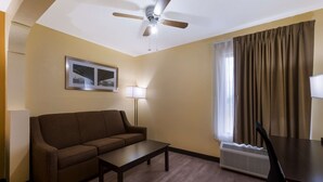 Habitación de hotel con un sofá marrón, una mesita, un ventilador de techo, una lámpara, una ventana con cortinas y un radiador.