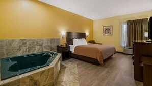 Habitación de hotel con bañera, cama con cubrecama marrón, mesita de noche con lámpara y un cuadro colgado en la pared.