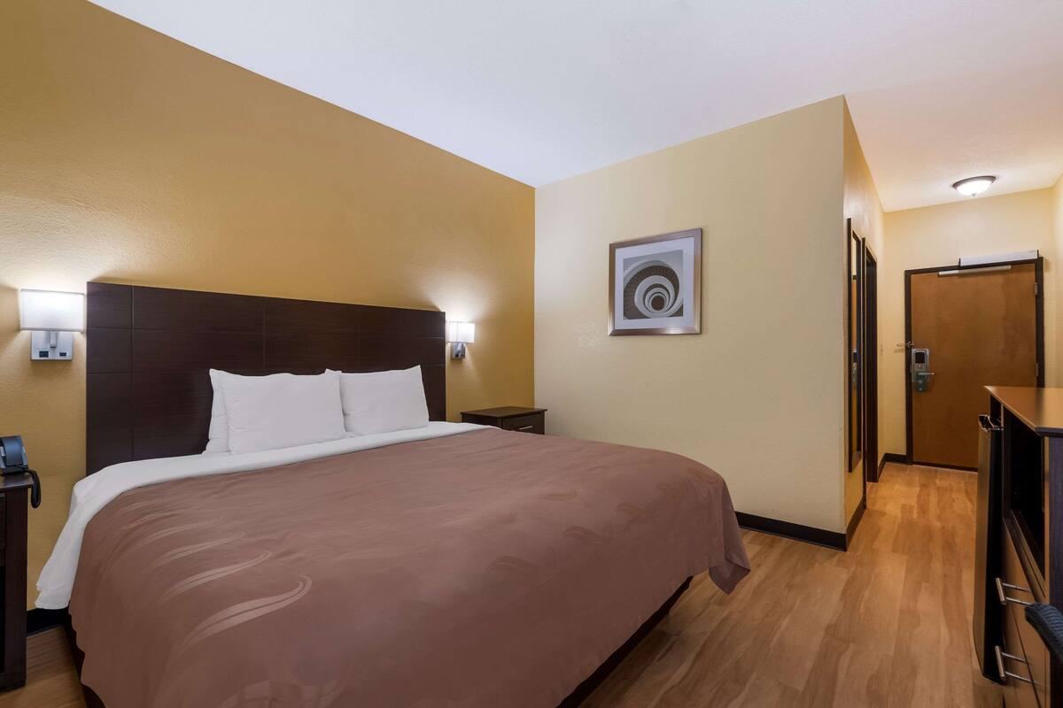 A hotel room with a large bed, bedside tables, a nightstand, a wall-mounted light, a door, and a wooden floor.