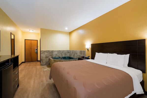 Habitación de hotel con una cama grande, un aparador de madera, un baño con bañera y una puerta que da a otra habitación.