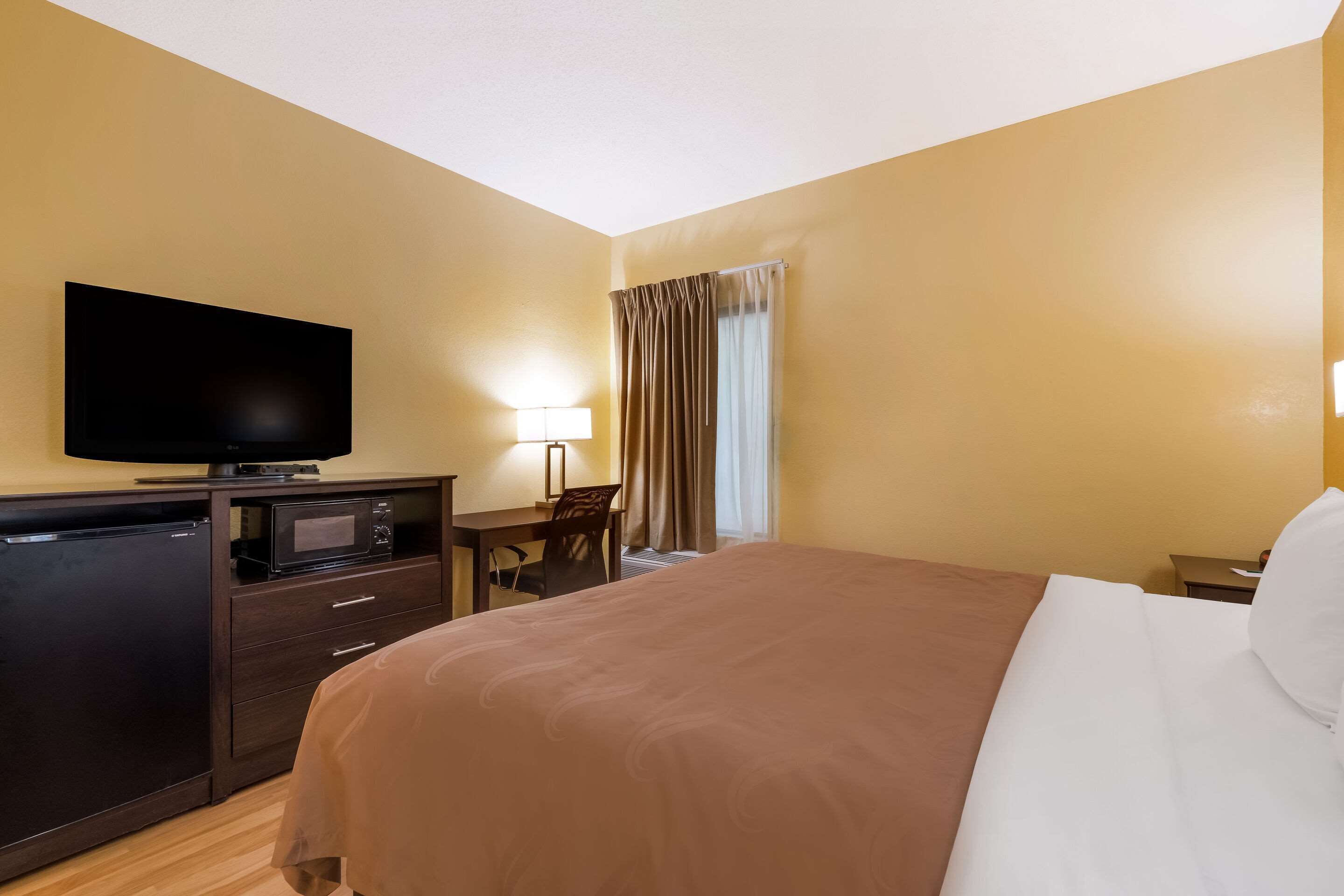 A hotel room with a bed, a desk with a lamp, a television, and a mini-fridge.