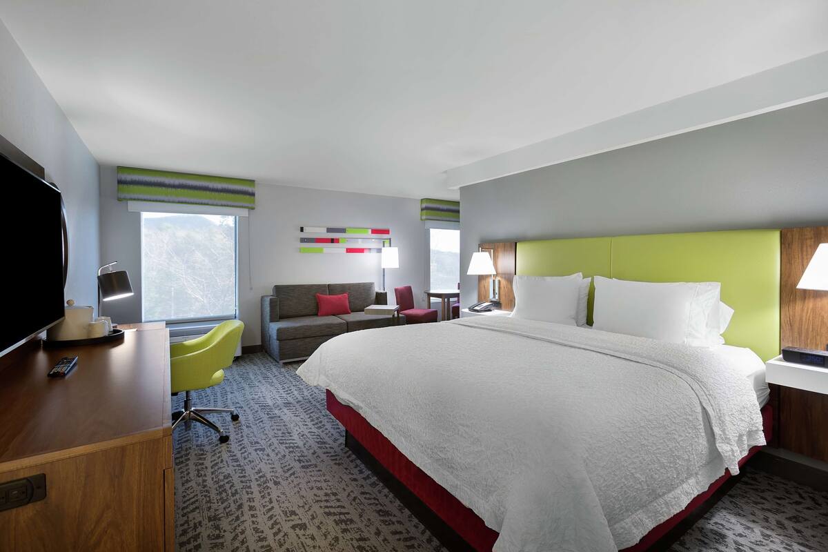 Ein Hotelzimmer mit einem großen Bett, einem Schreibtisch mit Fernseher, einer Couch, einem Sessel und einem Fenster mit Bergblick.