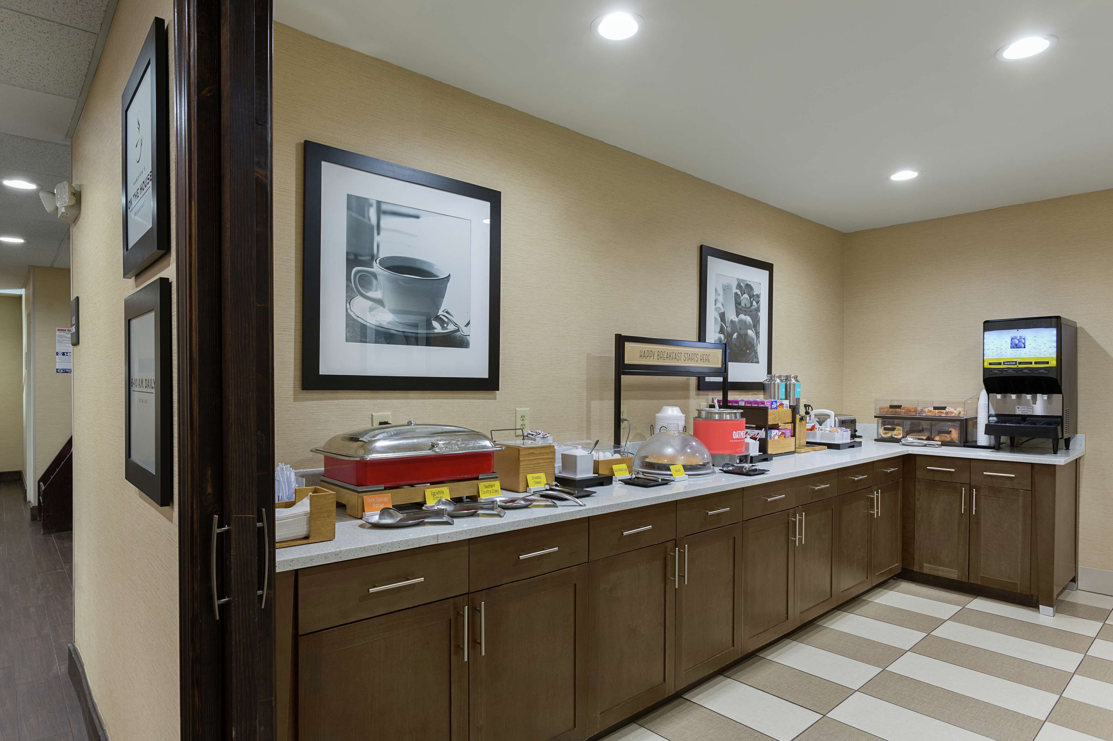 A hotel kitchen with a buffet setup, including a coffee machine, various food items, and framed pictures on the wall.