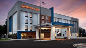 Et Hampton Inn hotel med moderne design, der viser et tydeligt amerikansk flag, en godt oplyst indgang og en parkeringsplads.