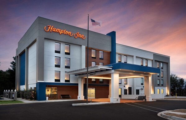 A Hampton Inn hotel with a modern design, featuring a prominent American flag, a well-lit entrance, and a parking area.