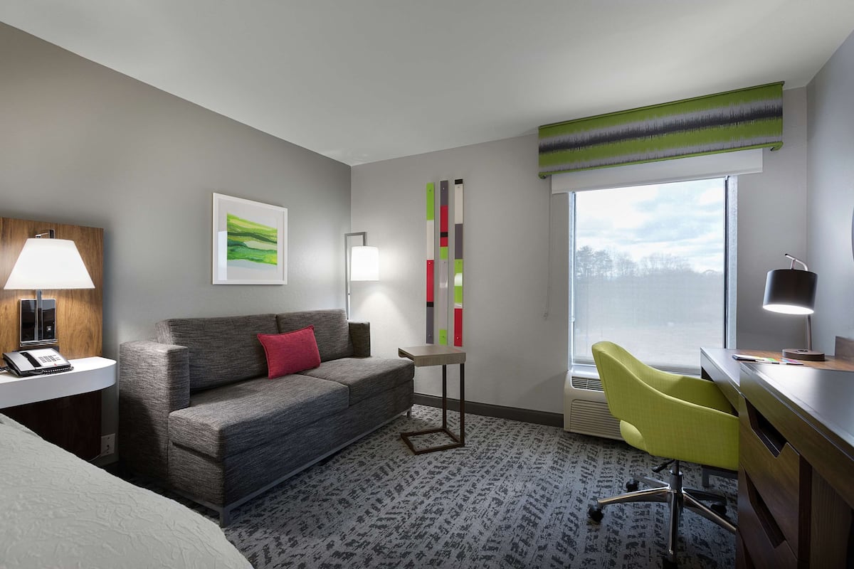 Ein Hotelzimmer mit grauem Sofa, einem kleinen Tisch, einem Schreibtisch mit Lampe, einem grünen Sessel, einem Fenster mit Aussicht und einer Wand mit vertikalem Farbdesign.