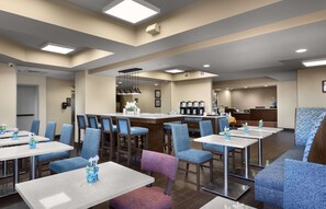 A hotel lobby with multiple seating areas, including tables and chairs, a reception desk, and a coffee station.