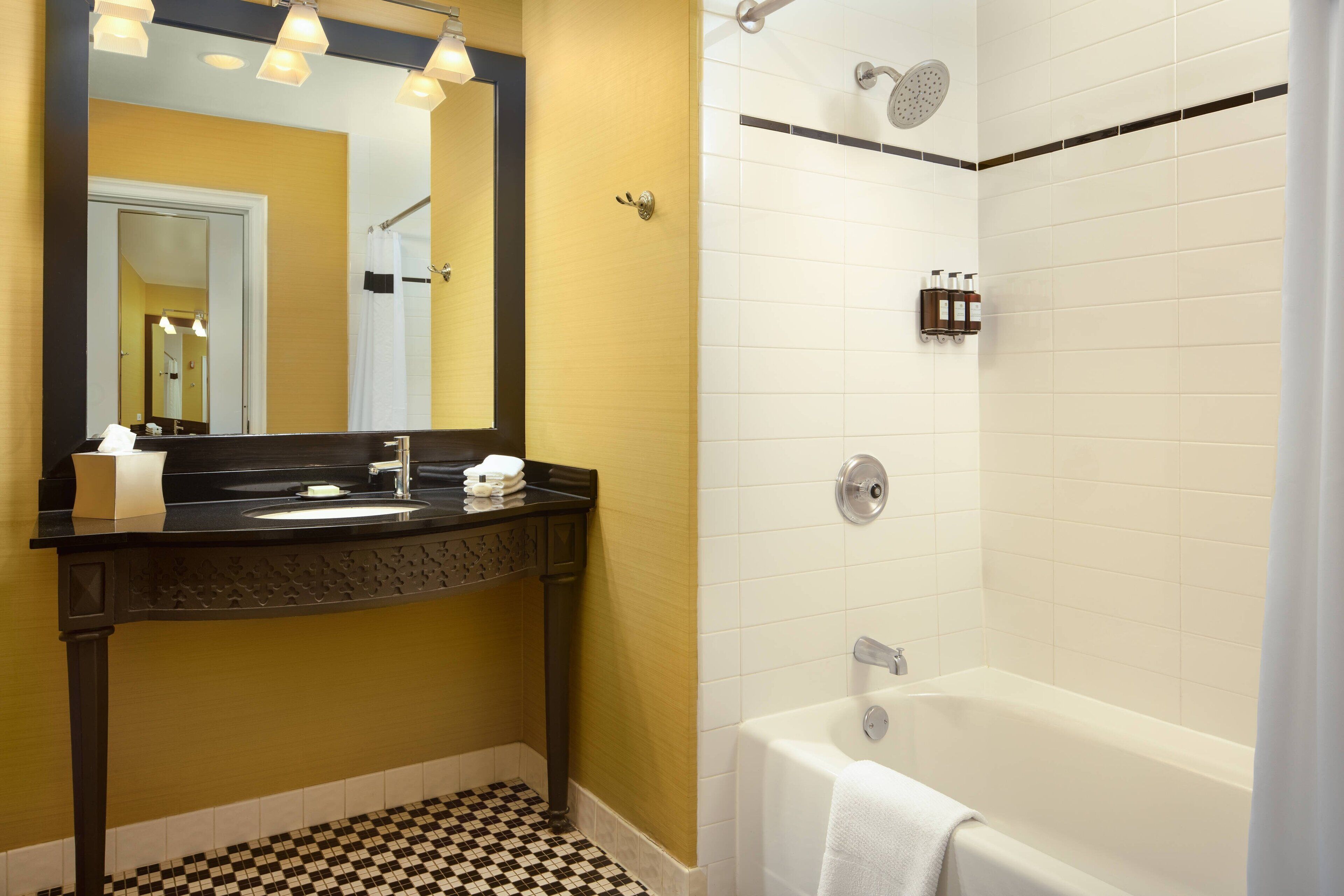 A bathroom with a black and white checkered floor, a black vanity with a mirror, a white bathtub, and a shower with a rain showerhead.