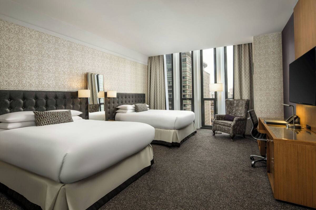 A hotel room with two beds, a desk, a chair, and a TV.