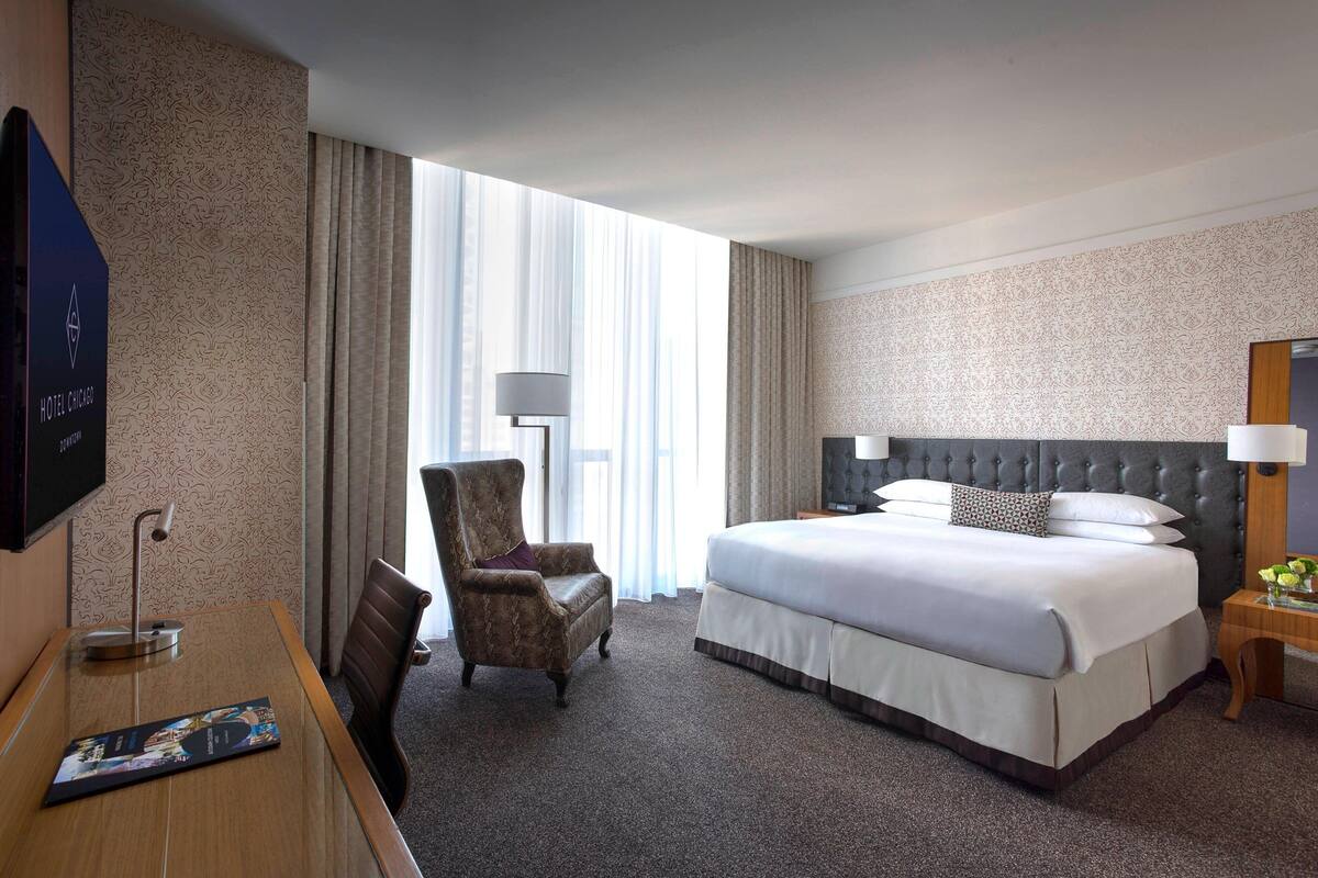 A hotel room with a large bed, a chair, a desk with a lamp, and a TV on the wall.