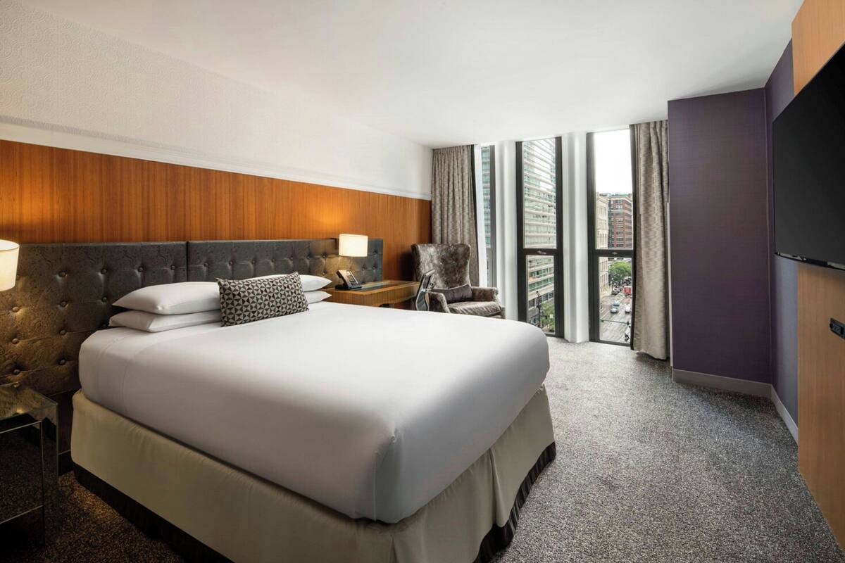 A hotel room with a large bed, a chair, a nightstand, a TV, and a view of the city through large windows.