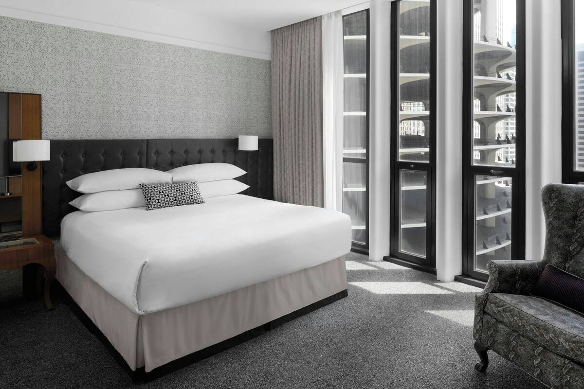A hotel room with a large bed, a nightstand, a chair, and a view of the building's interior through a glass door.
