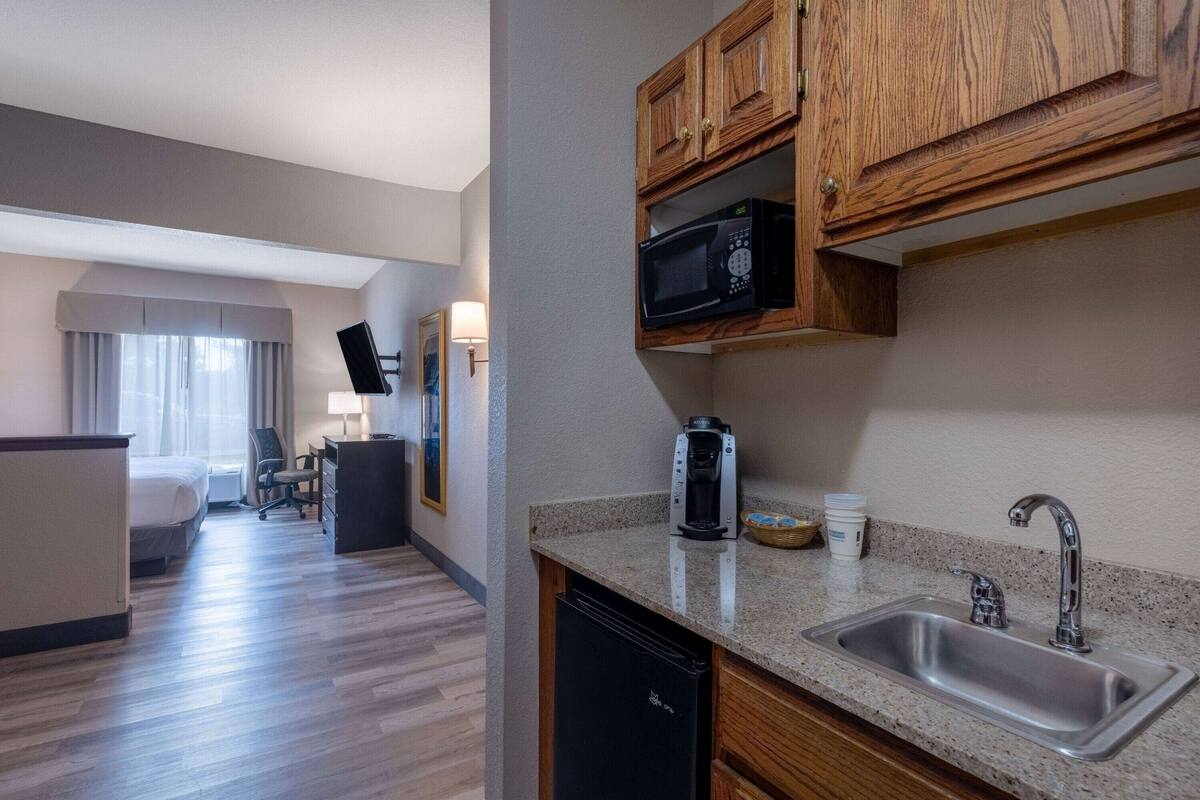 A hotel room with a kitchenette, including a microwave, coffee maker, and sink. There is a bed, a desk, and a chair in the room.