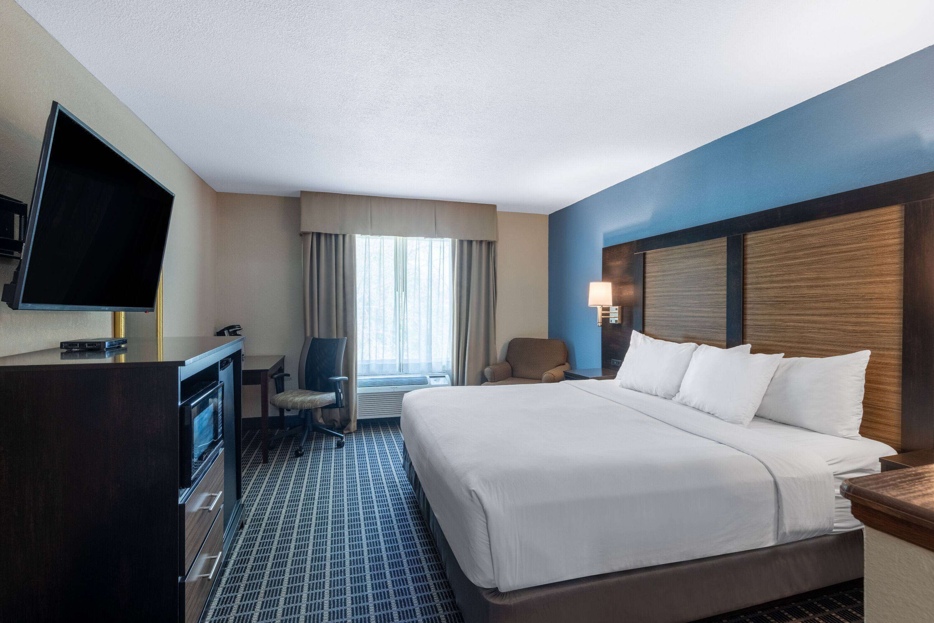 A hotel room with a large bed, a desk with a flat-screen TV, a chair, and a window with curtains.
