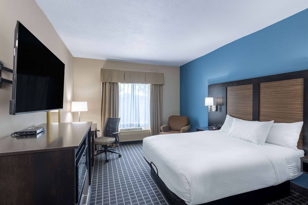 A hotel room with a large bed, a desk with a flat-screen TV, a chair, a window with curtains, and a nightstand with a lamp.