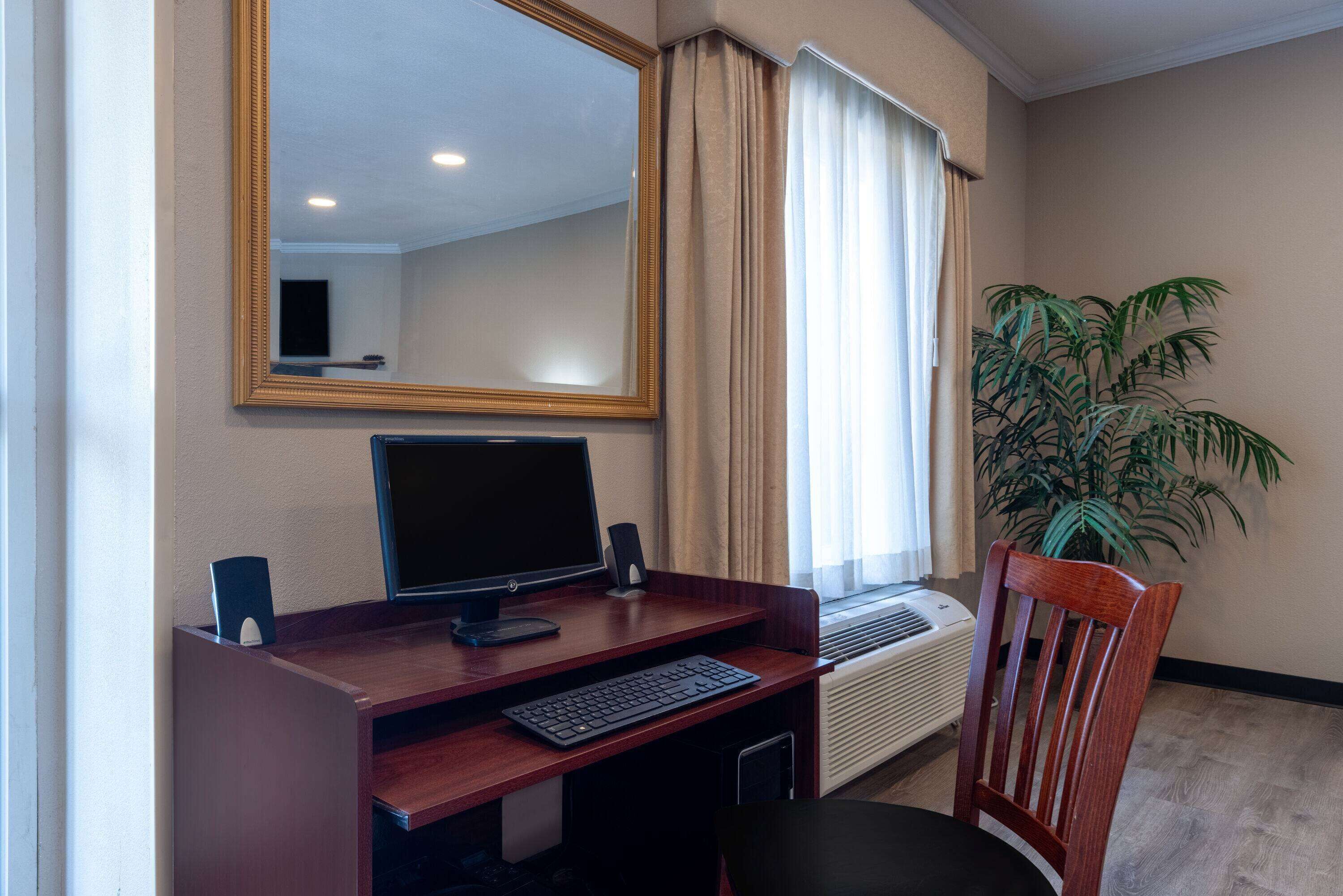A hotel room with a wooden desk, a computer, a chair, a large mirror, a potted plant, and a window with curtains.