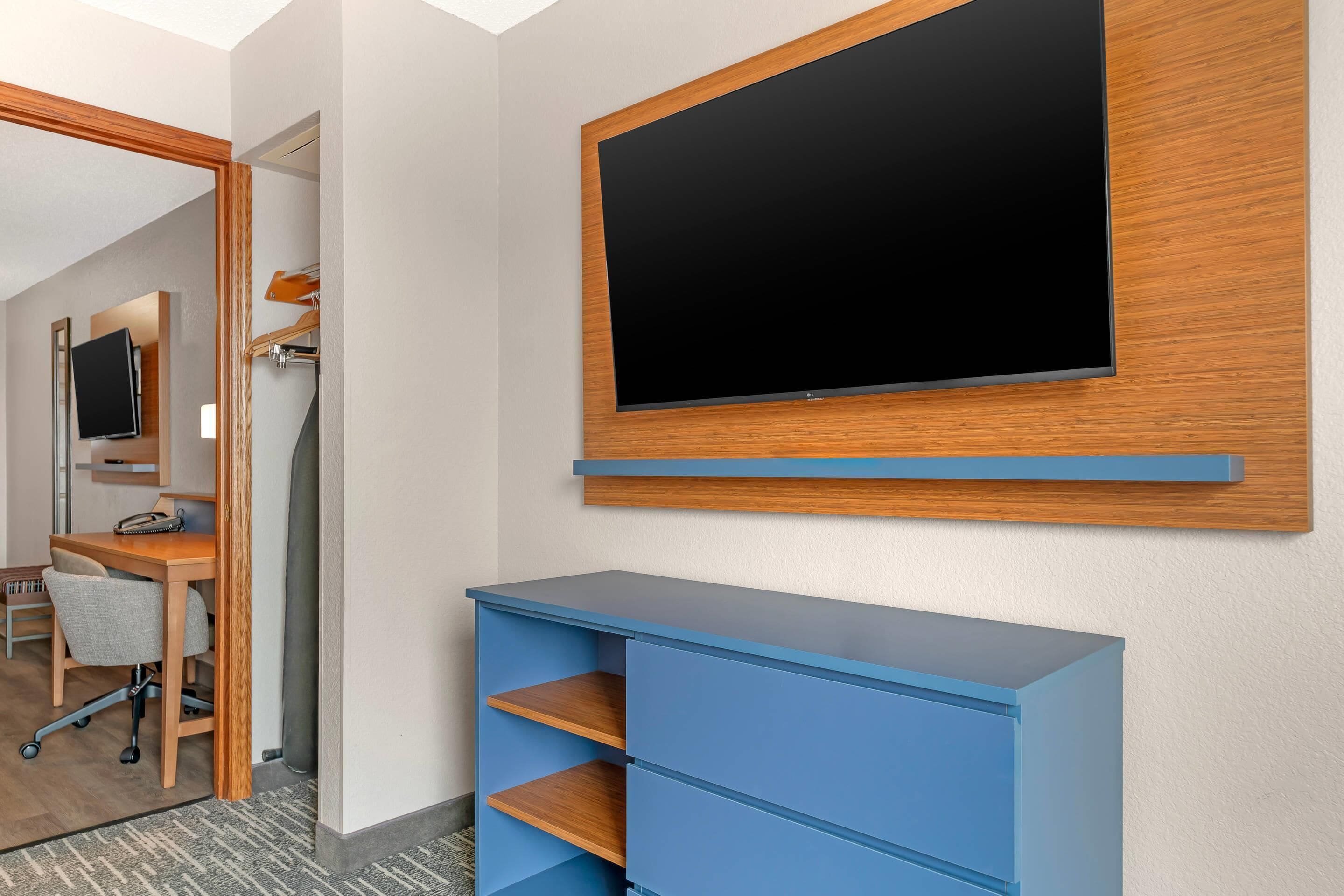 Ein Hotelzimmer mit flachbildfernseher an der Wand, einem Schreibtisch mit Stuhl und einer blauen Kommode.
