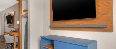 Ein Hotelzimmer mit flachbildfernseher an der Wand, einem Schreibtisch mit Stuhl und einer blauen Kommode.