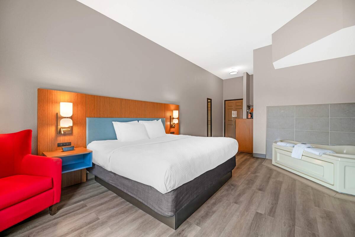 A modern hotel room with a large bed, a red armchair, a nightstand with a lamp, and a bathtub.