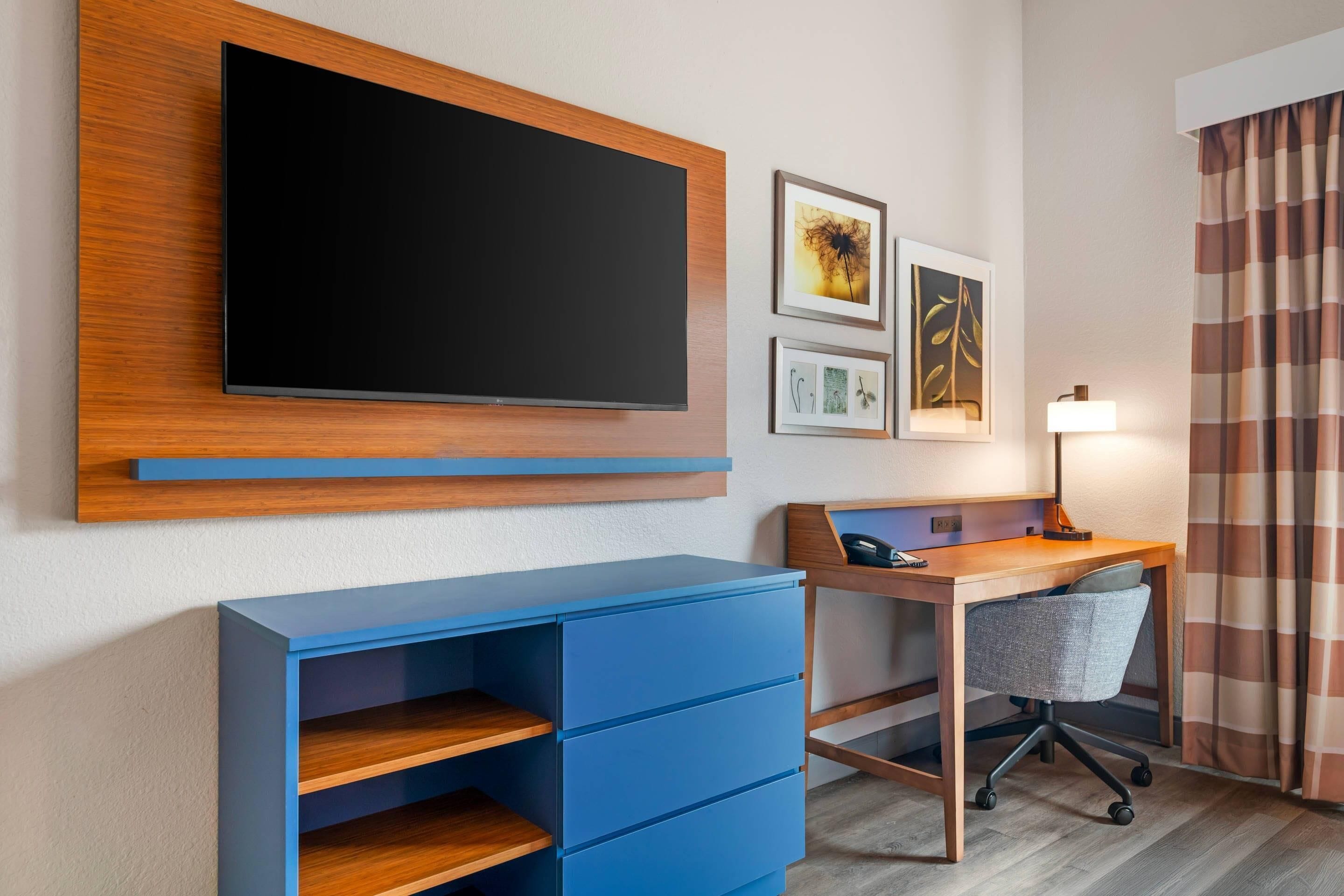 Et hotelværelse med et fladskærms-tv på væggen, et skrivebord med stol og et blått opbevaringsskab.