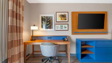 A hotel room with a wooden desk, a blue storage unit, a grey office chair, a lamp, and framed pictures on the wall.