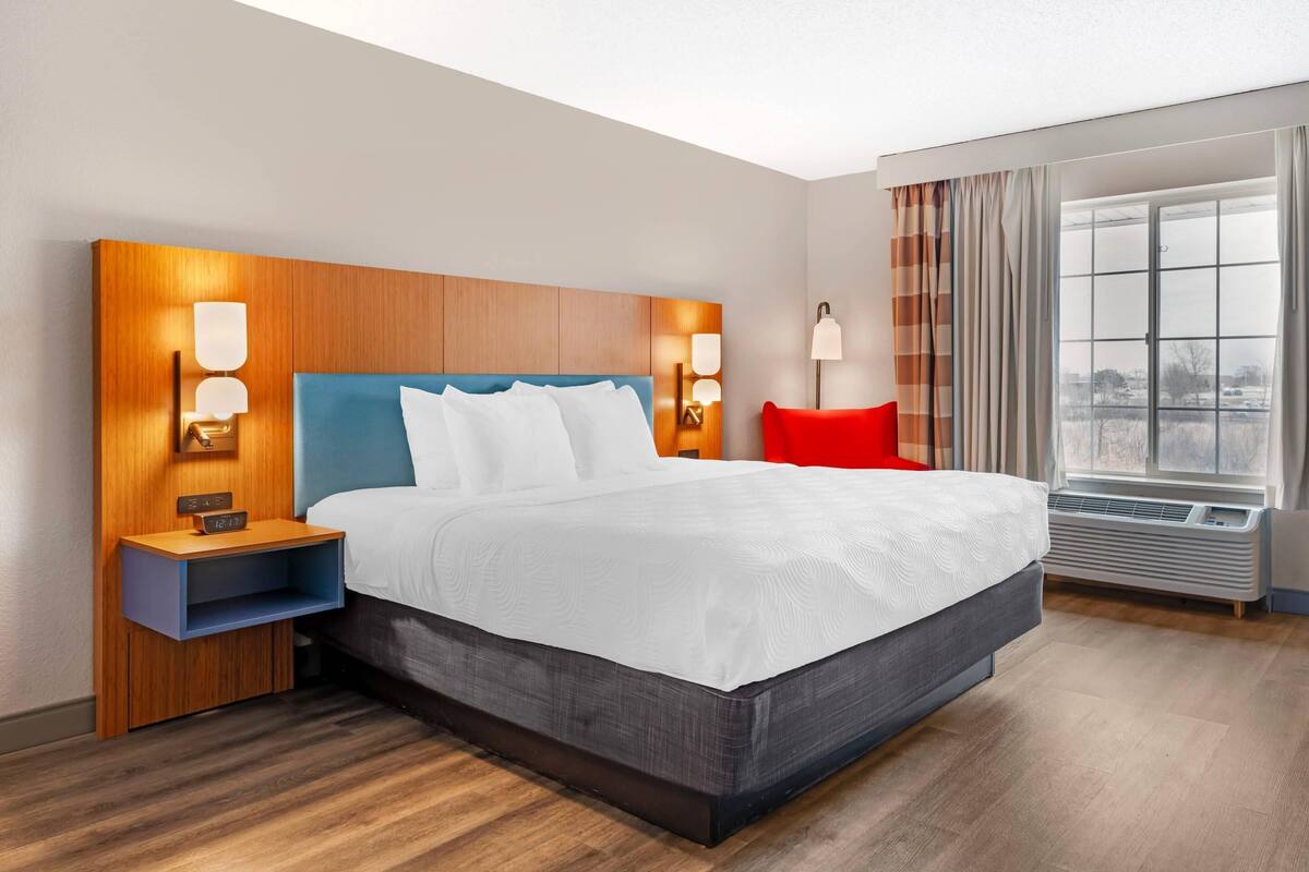 A hotel room with a bed, nightstands, wall-mounted lamps, a window with curtains, and an air conditioning unit.