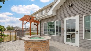 A patio area with a stone fire pit, a wooden pergola, and a house with large windows and a door.
