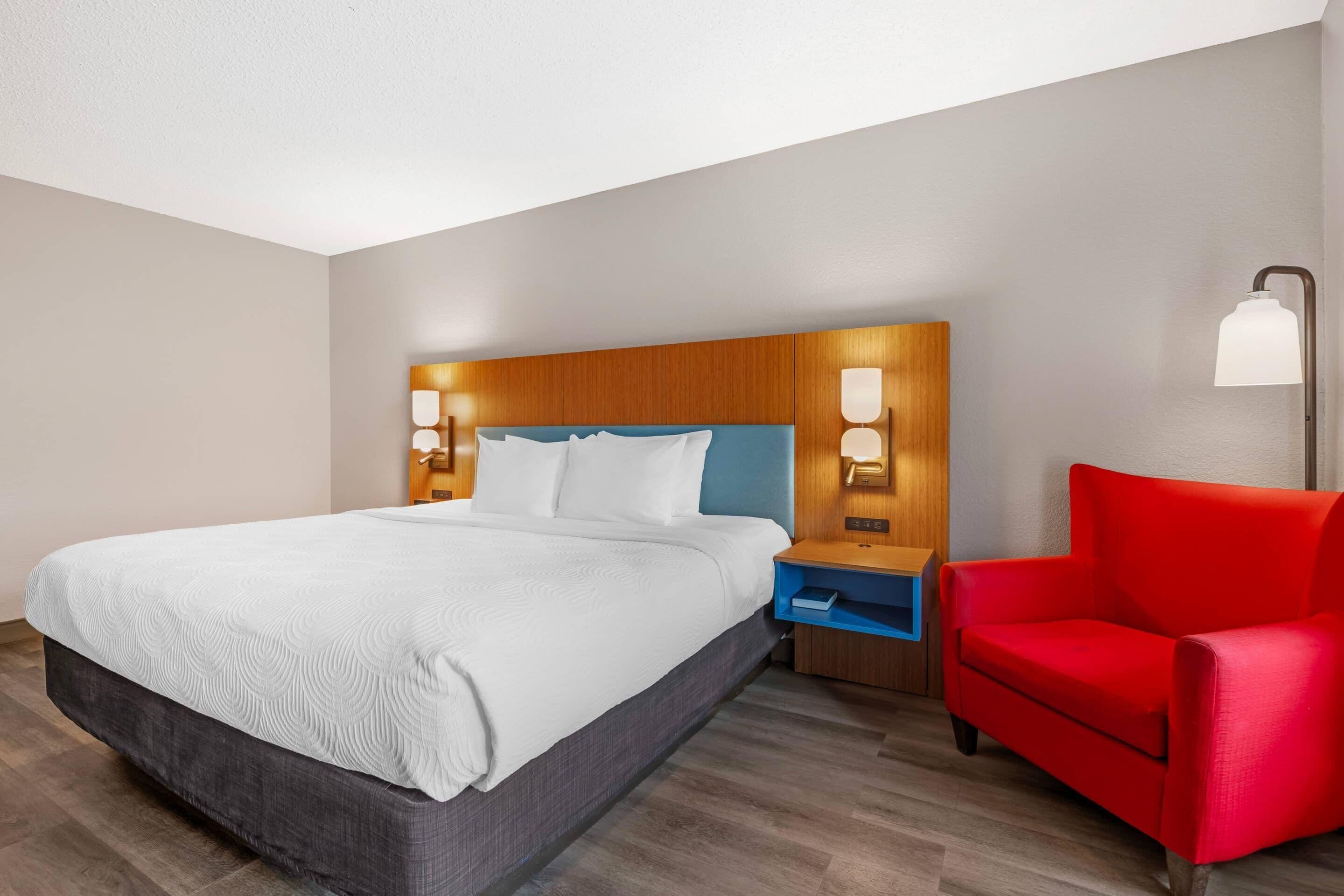 Una habitación de hotel con una cama grande, cabecera de madera, mesitas de noche con lámparas, un sillón rojo y una mesita auxiliar azul.