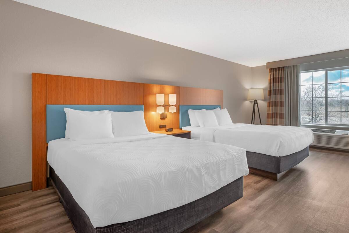 A hotel room with two beds, a wooden headboard, a nightstand with a lamp, and a large window with a view.