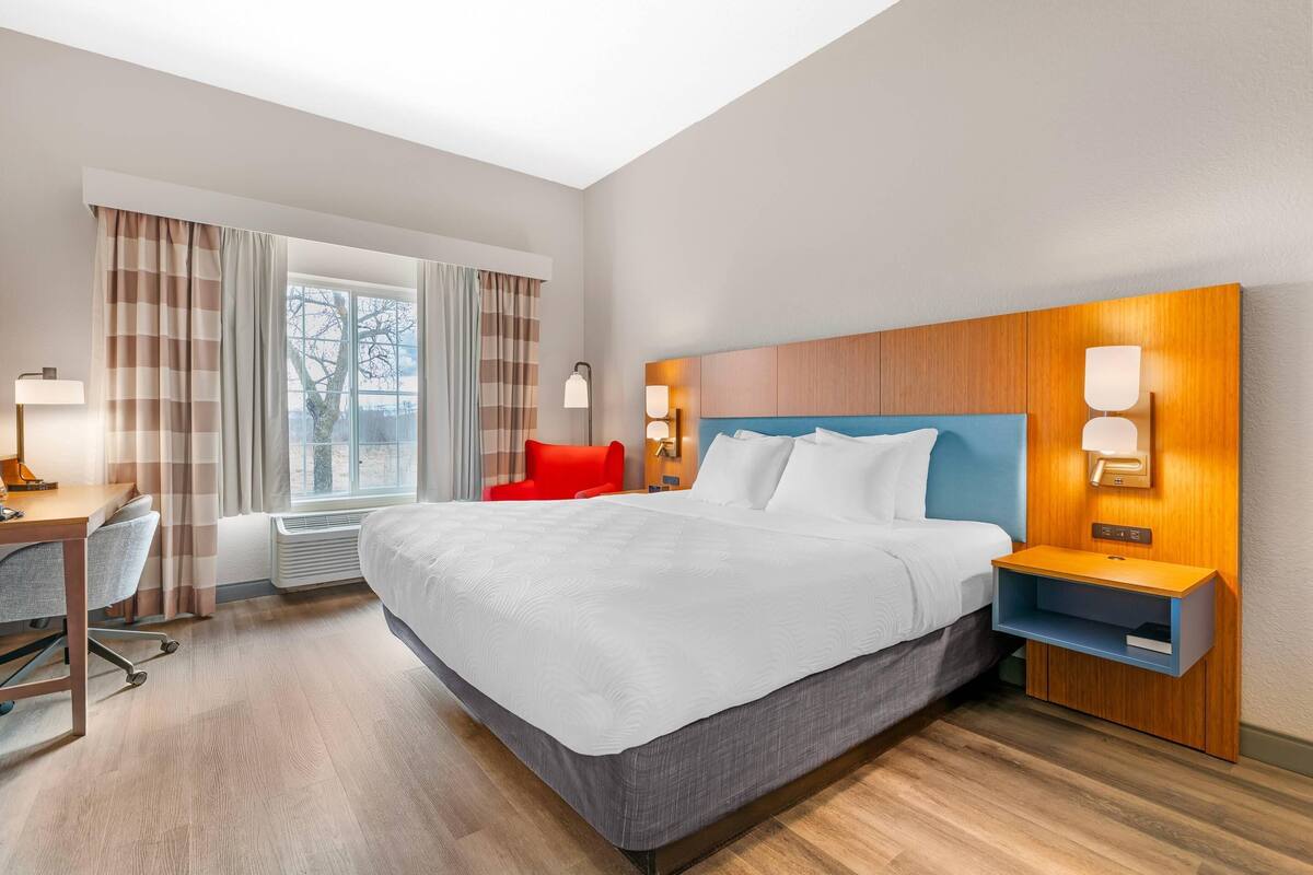 A hotel room with a large bed, a desk, a chair, a nightstand, and a window with curtains.