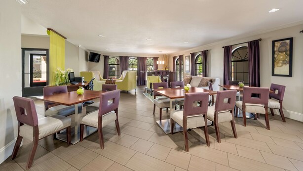 A spacious dining area with a large wooden table and matching chairs, a kitchen in the background, and a television mounted on the wall. - SureStay Hotel by Best Western Deer Park (Deer Park)