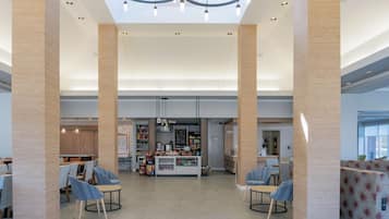 A spacious lobby with high ceilings, a central seating area with round tables and chairs, and a food kiosk in the background.