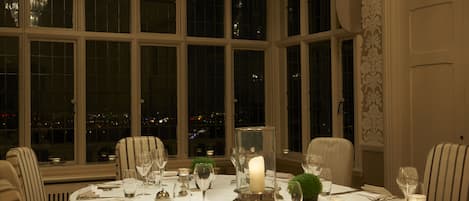 A dining table set for a meal with chairs, a candle centerpiece, and a view of the city at night through large windows.