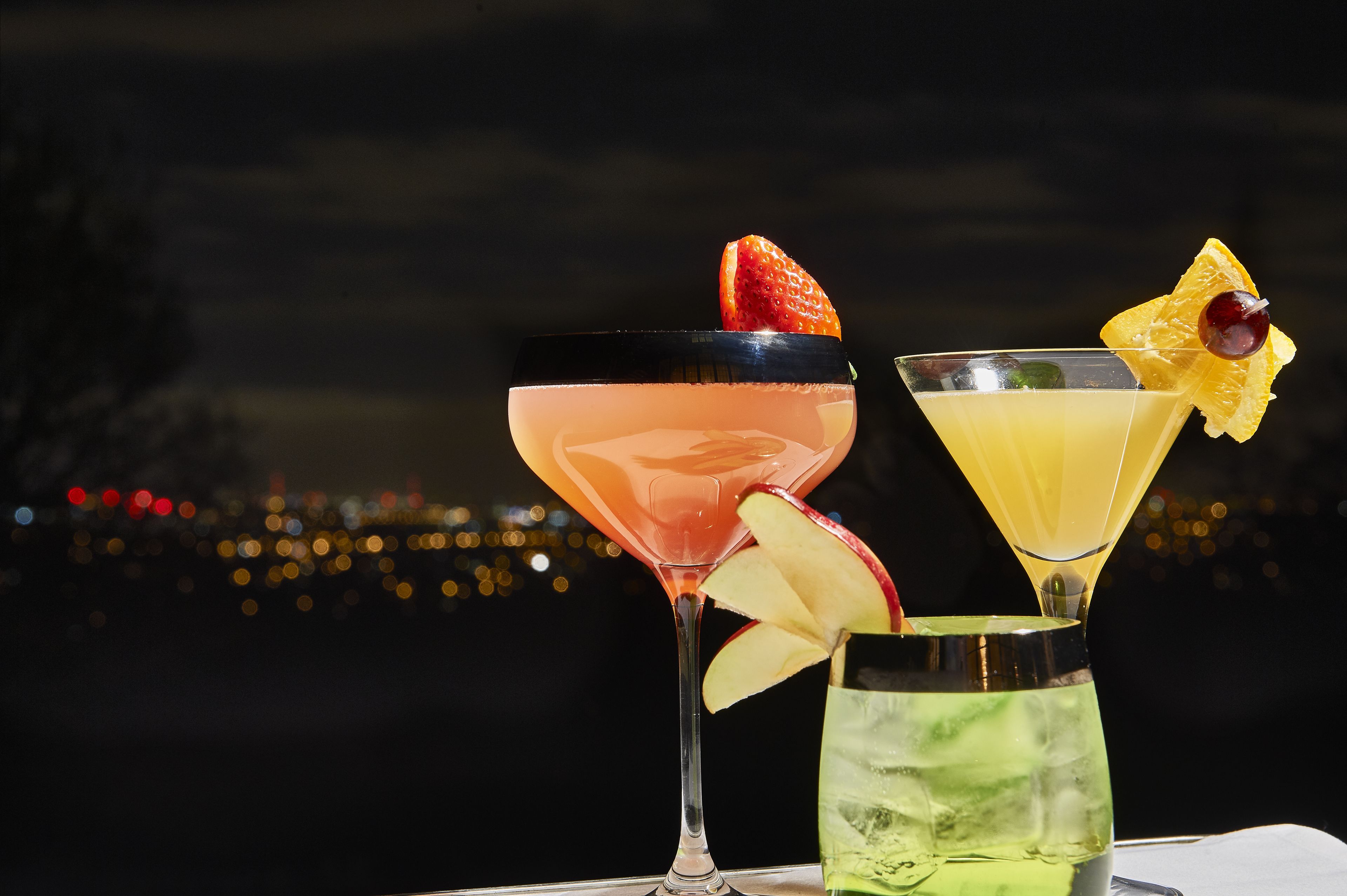 Three cocktails with fruit garnishes on a table with a cityscape view in the background.