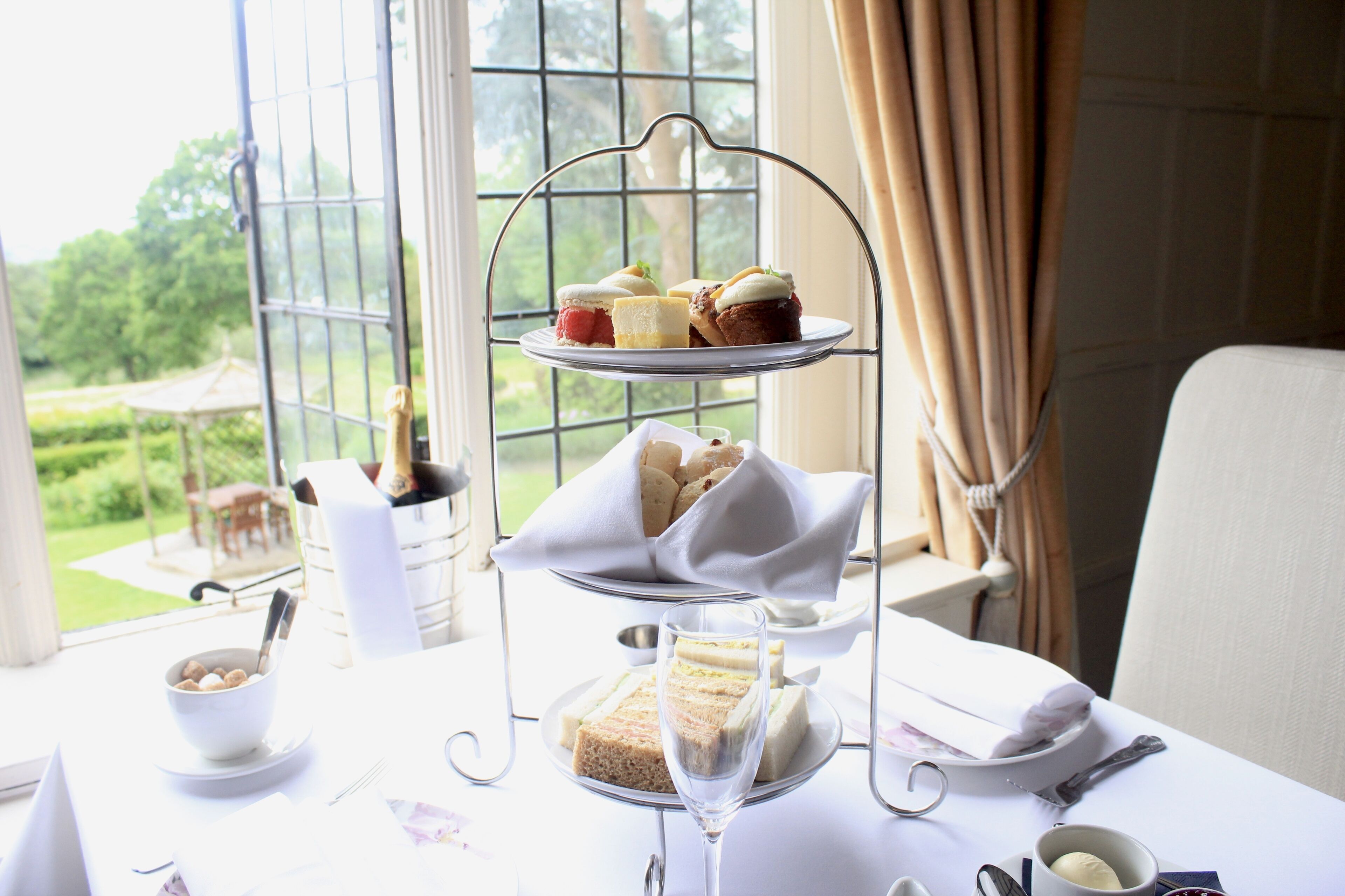 A three-tiered dessert stand with sandwiches and cakes, a glass of champagne, and a bowl of cookies on a table with a view of a garden through a window.