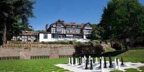 A large, traditional Tudor-style building with a well-maintained lawn and a chessboard in the foreground. - The Manor Elstree (Borehamwood)