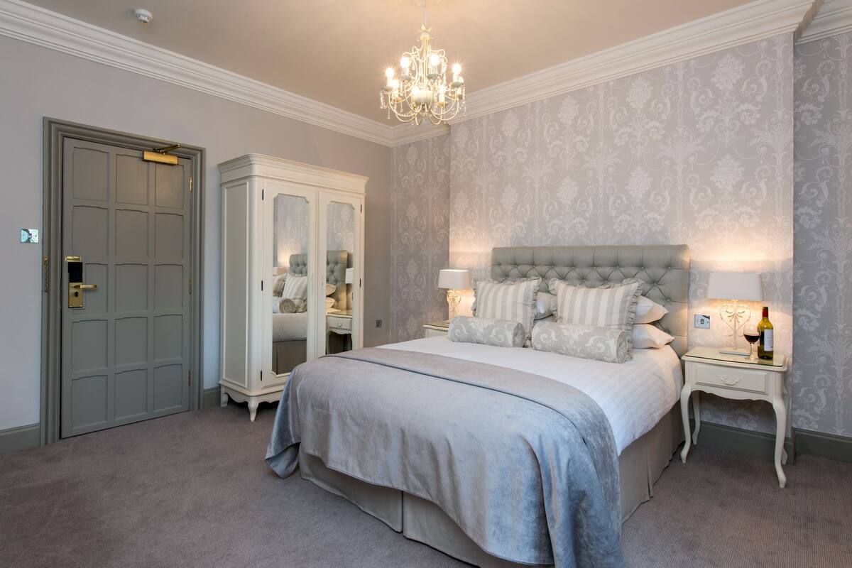 A bedroom with a large bed, a wardrobe, a chandelier, and a nightstand with a lamp and a bottle.