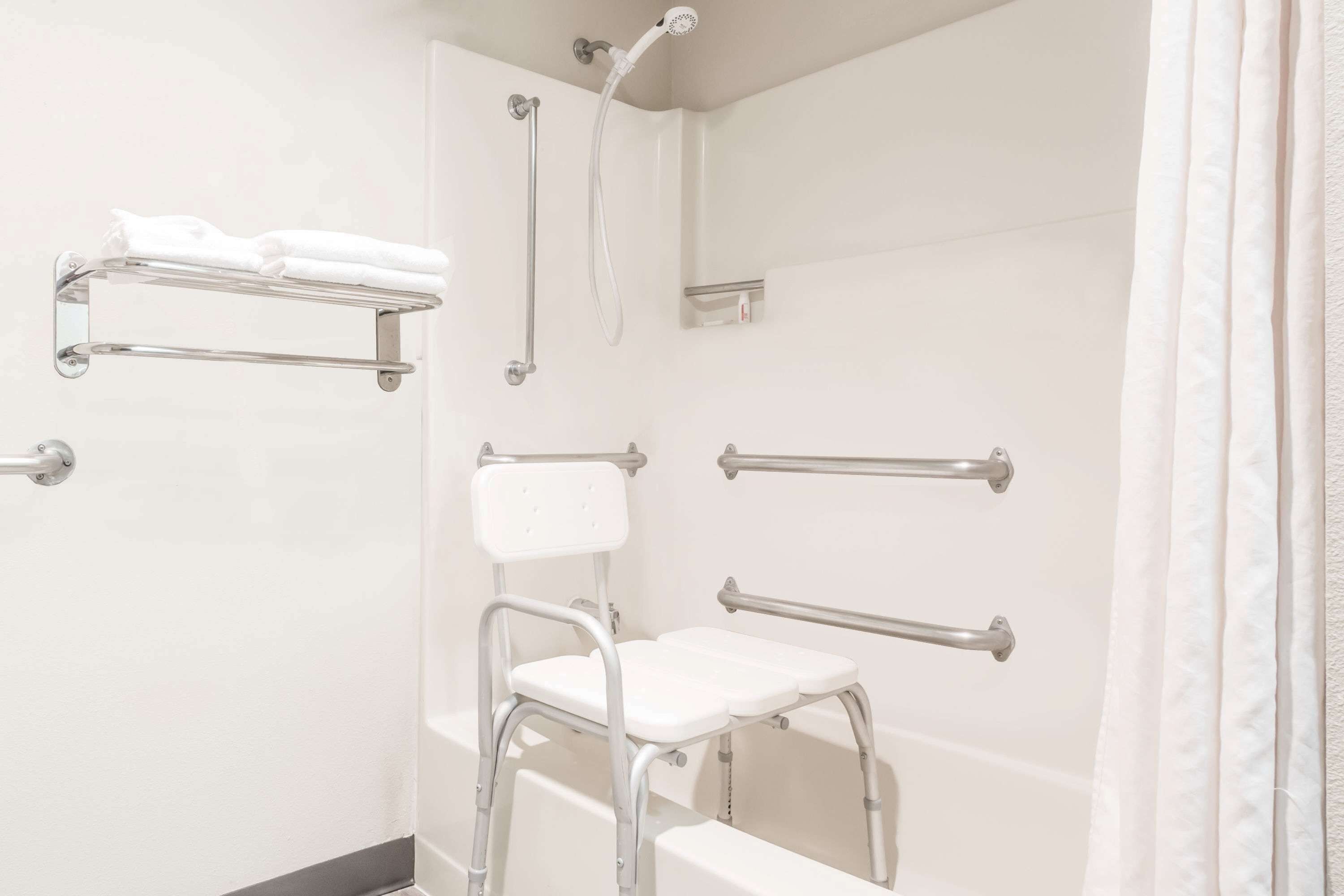 A bathroom with a shower, grab bars, a white chair, and a towel rack.